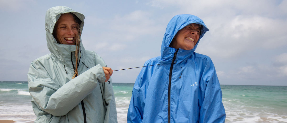 Two people wearing lightweight hooded rain jackets - Allweather Coat stand on a beach, laughing while one pulls on the drawcord of the other jacket. The jacket on the left is a pale mint green and the jacket on the right is a medium blue.