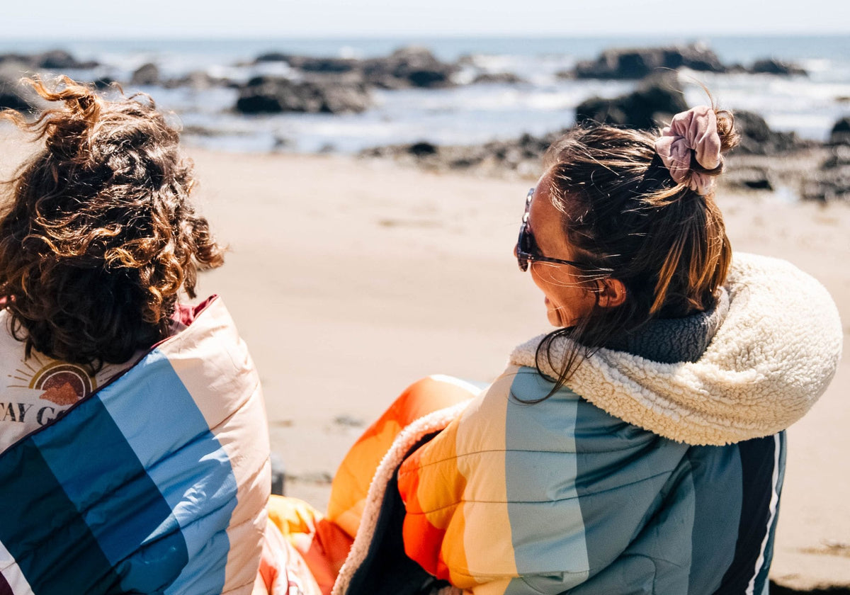 Two people sitting on a sandy beach facing the ocean, wrapped in colorful Cloudtouch blankets VOITED with blue, orange, cream, and gray panels.
