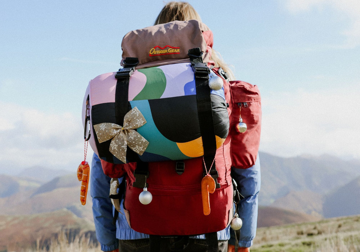 A person viewed from behind wearing a large, highly decorated hiking backpack on a mountain trail. Cloudtouch VOITED Blanket.