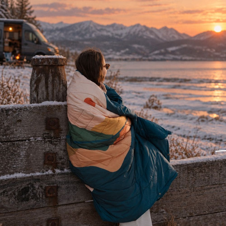 Woman wrapped in a VOITED Cloudtouch blanket with a colorful peach, mustard and green pattern, looking out over a snowy lake at sunset. AI modified
