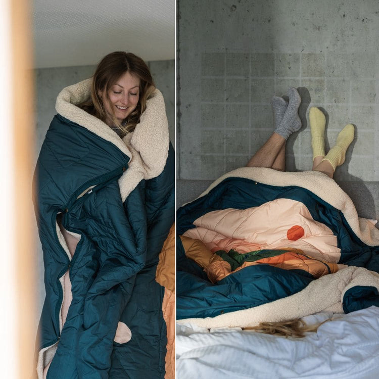 Woman wrapped in a VOITED Cloudtouch  QUEEN blanket with cozy sherpa lining; two people lying under the blanket on a bed with their socked feet up against a concrete wall.